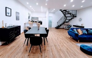 A modern living space with a dining area, a blue sofa, a staircase, and a kitchen in the background.