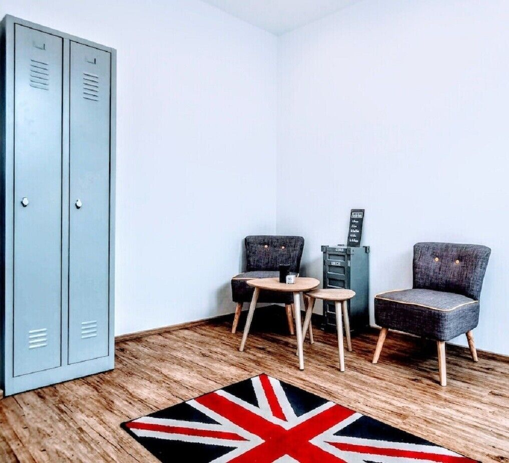 A corner room with a metal locker, two chairs, a small table, and a Union Jack rug.