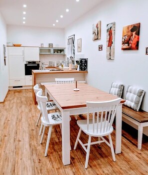 Una cocina moderna con armarios blancos, una mesa de comedor de madera y sillas blancas. En la zona de estar, hay un sofá con cojines a rayas.