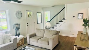 Un salon avec un canapé beige, une table basse, une horloge, un escalier et une porte dotée de panneaux de verre.