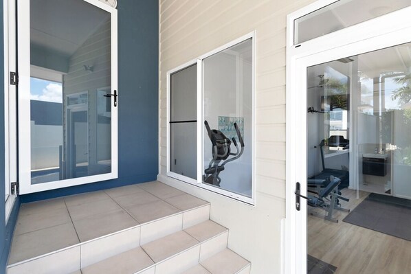 Un intérieur moderne avec un escalier menant à une porte vitrée, un appareil de musculation visible et un aperçu d’un espace de vie avec un téléviseur.