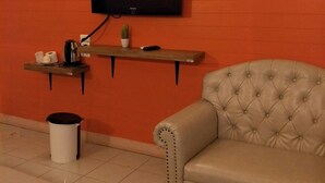 Ein Zimmer mit einem an einer orangefarbenen Wand montierten Fernseher, einem Sofa, einem kleinen Tisch mit einer Pflanze und einem Mülleimer.