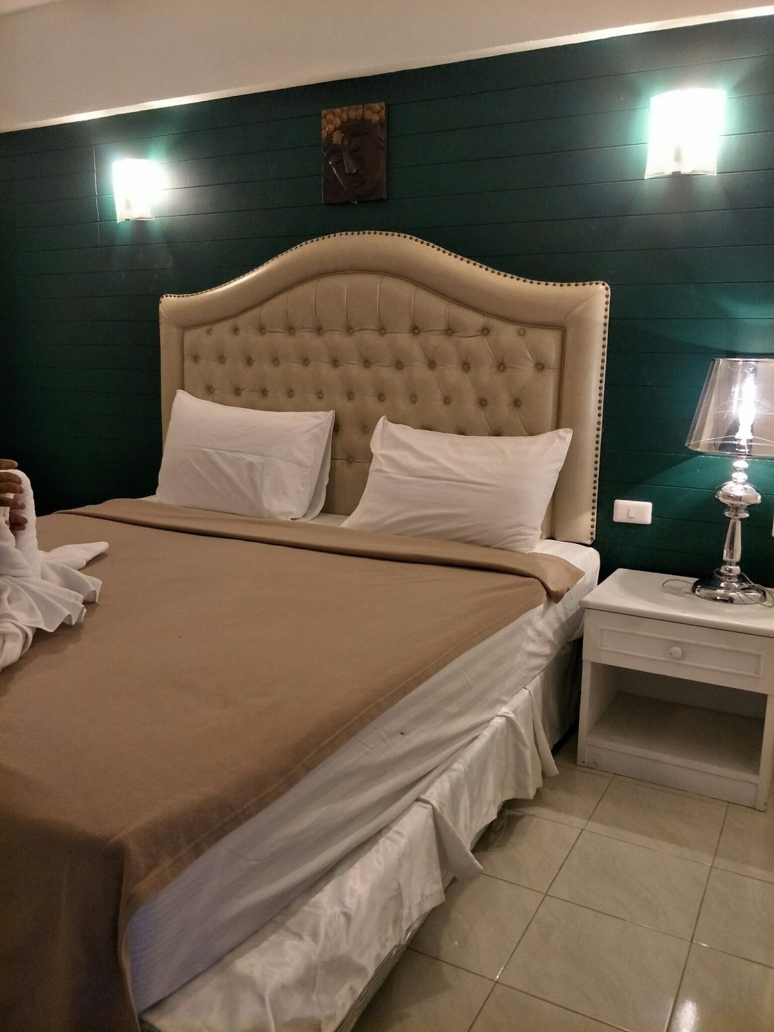 A neatly made bed with a tufted headboard, white pillows, and a brown bedspread. There is a white nightstand with a lamp and a small drawer. The room has dark green walls and a small painting above the bed.