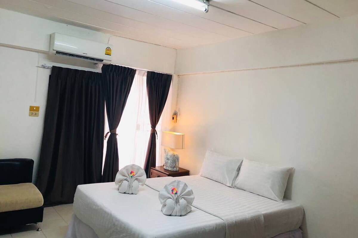 A hotel room with a bed, a chair, a nightstand, an air conditioner, and a window with curtains.