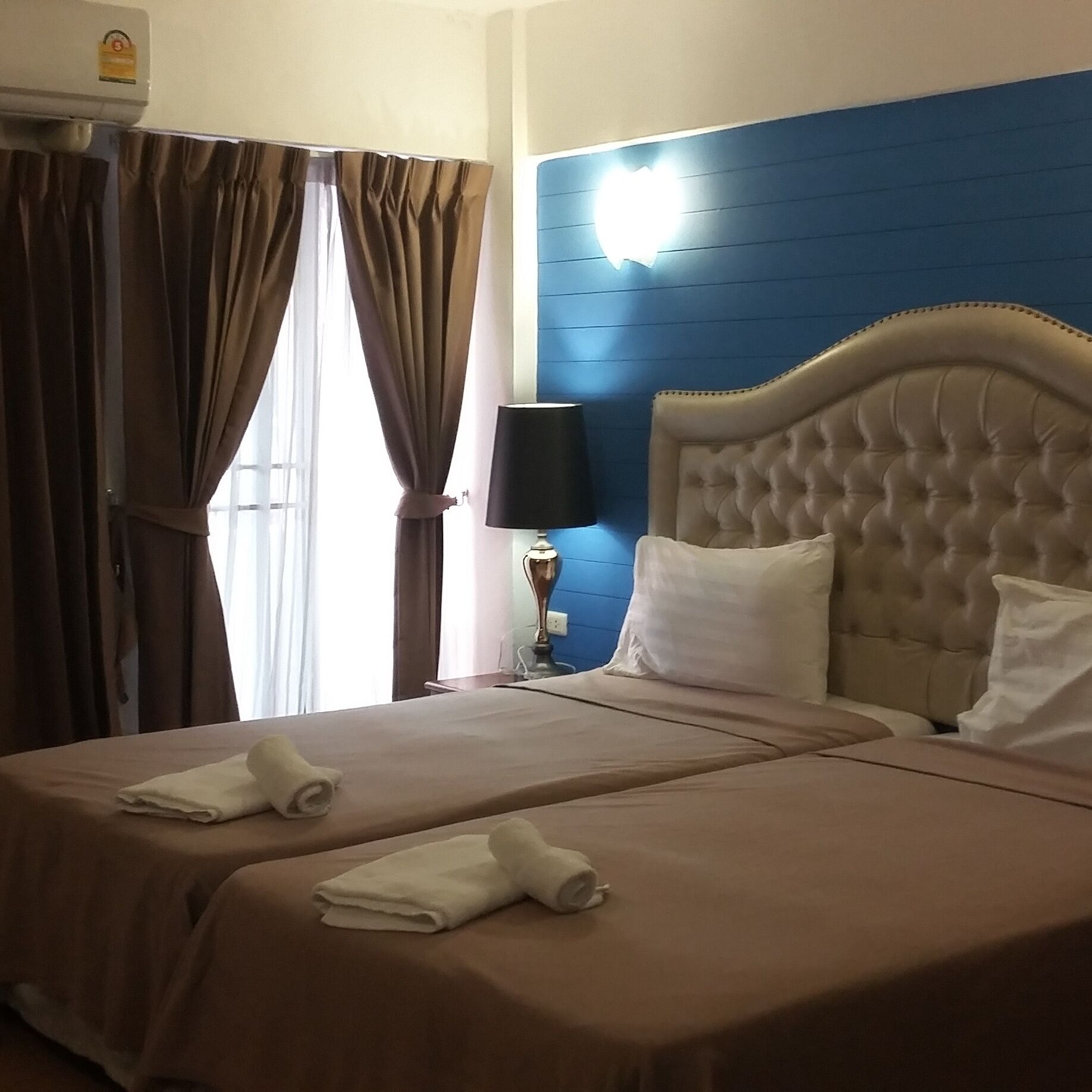 A hotel room with two beds, a blue accent wall, a window with curtains, and a lamp on a bedside table.