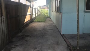 A narrow corridor with a concrete floor, wooden fence on one side, and a light blue building on the other. - Camping with a Roof, bathroom and Kitchen (Palm Beach)