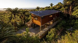 A wooden cabin with a deck, surrounded by lush greenery and overlooking a coastal area. - Treescapes (Punakaiki)