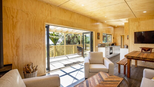 A spacious living area with a wooden ceiling, a large sliding glass door leading to a balcony, and a kitchen in the background. - Treescapes (Punakaiki)