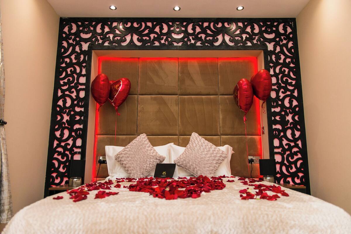 Een hotelkamer met een gestoffeerd hoofdeinde, decoratieve wandpanelen, en een bed versierd met rozenblaadjes en hartvormige ballonnen.