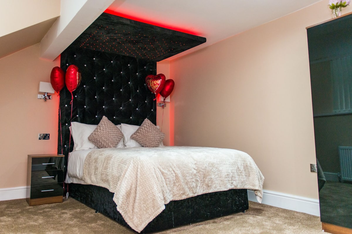 Een slaapkamer met een zwart hoofdeinde, een bed met wit beddengoed en rode hartvormige ballonnen.