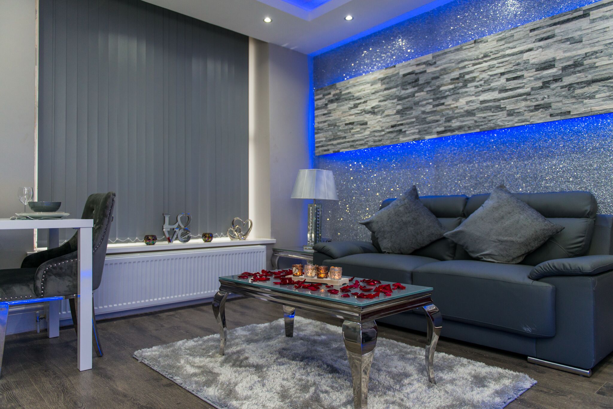 A modern living room with a grey sofa, a glass coffee table with rose petals, a white table with a bowl and glasses, and a decorative wall with a blue light strip.