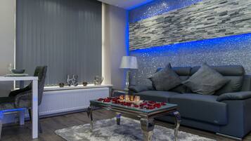 A modern living room with a grey sofa, a glass coffee table with rose petals, a white table with a bowl and glasses, and a decorative wall with a blue light strip.