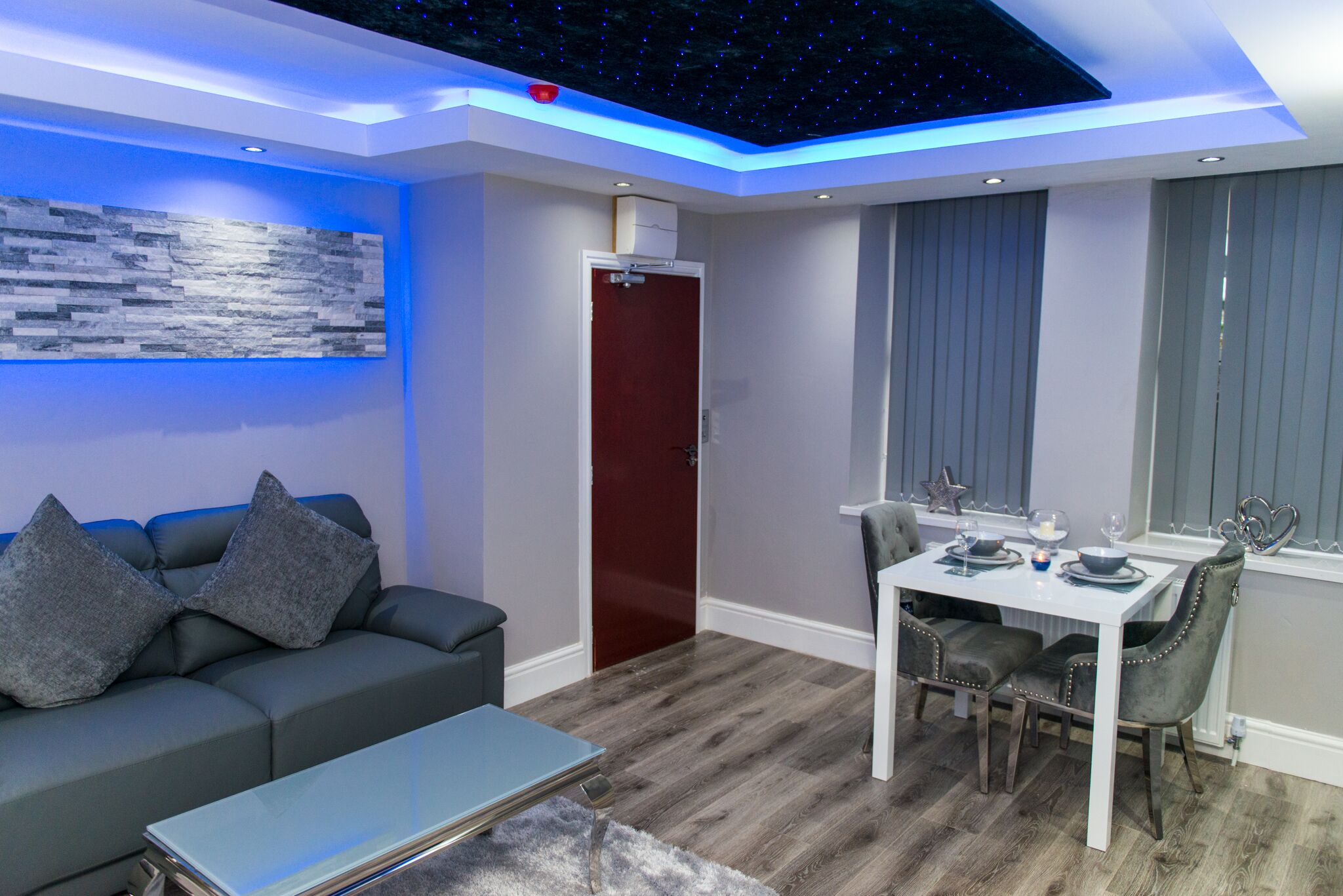 A modern living room with a grey sofa, a glass coffee table, and a dining area with a white table and grey upholstered chairs.