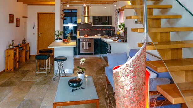 Un vaste espace de vie doté d’un plafond haut, de poutres apparentes, d’une cuisine équipée d’appareils modernes, d’un escalier et d’un coin repas avec une table et des chaises.