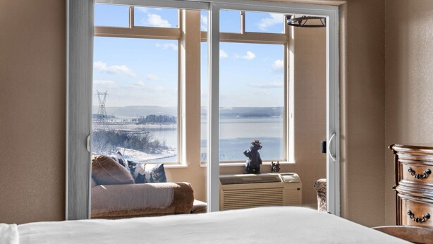 Ein Schlafzimmer mit großem Fenster und Blick auf einen See sowie Stromleitungen in der Ferne.