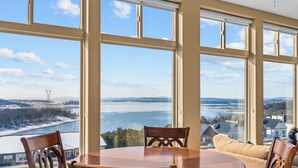 Ein Essbereich mit großem Tisch und Stühlen, große Fenster mit Blick auf ein Gewässer und entfernte Gebäude.