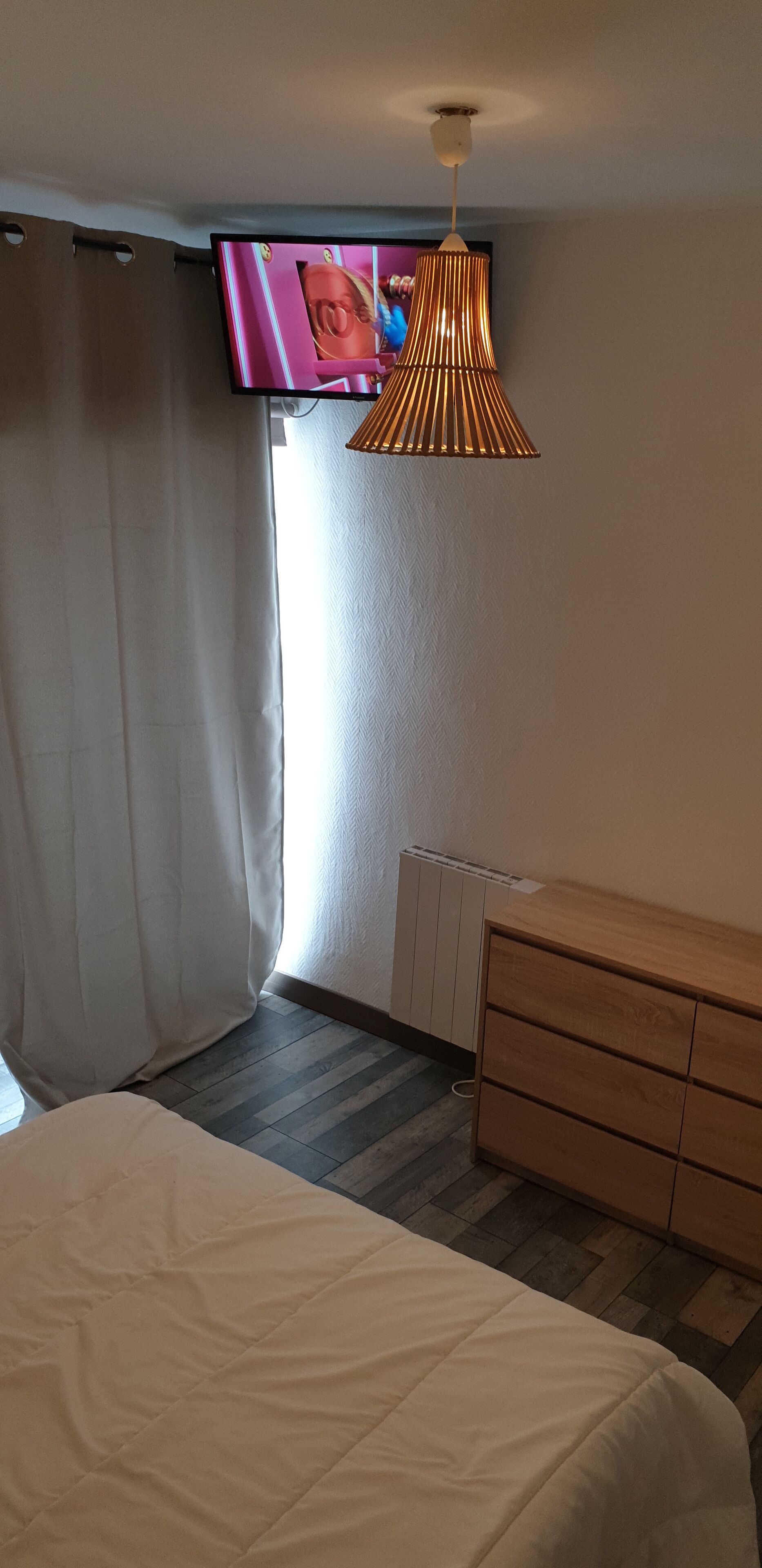 Une chambre d’hôtel avec un lit, une commode en bois, un téléviseur fixé au mur et un luminaire au plafond.
