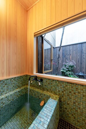 A wooden-paneled bathroom with a green-tiled bathtub, a window, and a view of a wooden fence and plants outside. - Yamagata Kyomachi Hatago Kyoto Station (Kyoto)