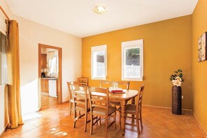 A dining area with a wooden table and chairs, a kitchen in the background, and a window with a view of greenery.