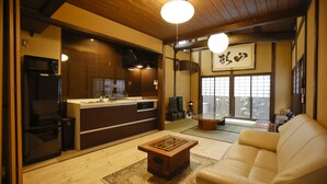 Un salon de style japonais traditionnel, comprenant un canapé, une table basse en bois, un coin cuisine avec un réfrigérateur et un four à micro-ondes, et une grande fenêtre donnant sur des tatamis.