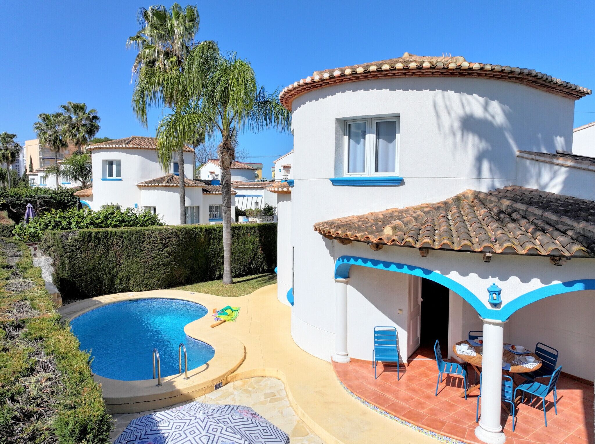 Ein Poolbereich mit einem gebogenen Pool, einer Terrasse mit Stühlen und Tischen sowie einem Gebäude mit einem gefliesen Dach und blauen Verzierungen.