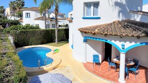 Un área de piscina con una pileta curva, un patio con sillas y mesas, y un edificio con techo de tejas y detalles azules.