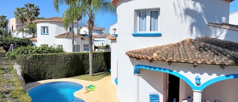 A pool area with a curved pool, a patio with chairs and tables, and a building with a tiled roof and blue trim.
