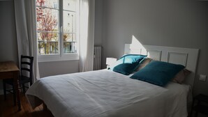 A neatly made bed with white bedding and two teal pillows, a wooden desk with a chair, and a window with a view of buildings and trees.