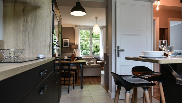 A modern kitchen with dark cabinetry, a dining area with a table and chairs, and a view of a green outdoor area through a window.
