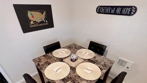 Ein Esstisch mit vier Platzsets, Stühlen, einer gerahmten Karte und einem Schild mit der Aufschrift 'TOGETHER WE ARE HOME'.