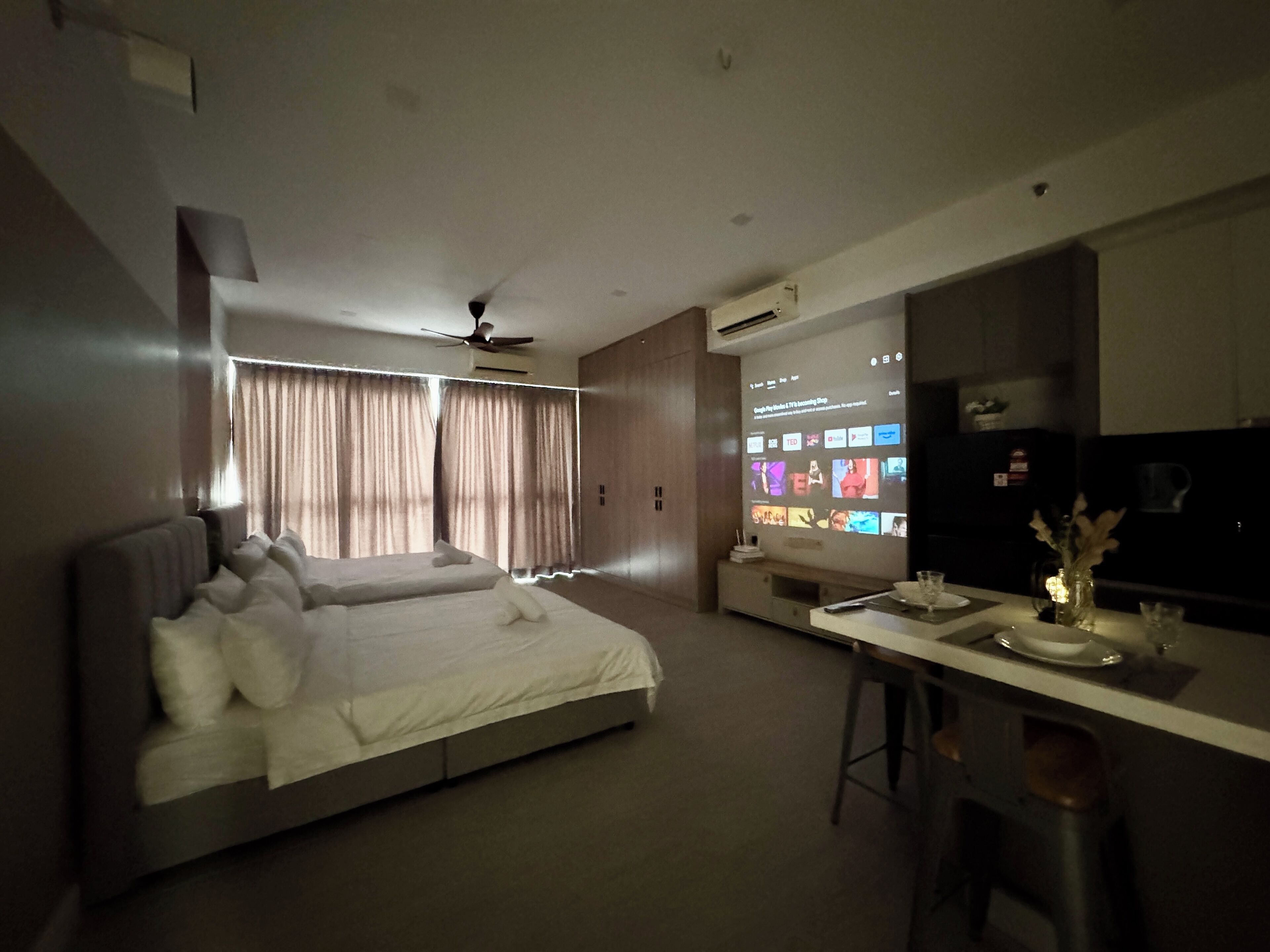 A modern hotel room with a large bed, a desk with a computer, and a kitchen area.