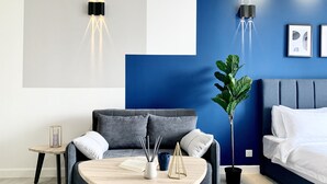 Une chambre à coucher moderne avec un canapé gris, une table ronde en bois, une plante sur la table de chevet et un lit avec une literie blanche.