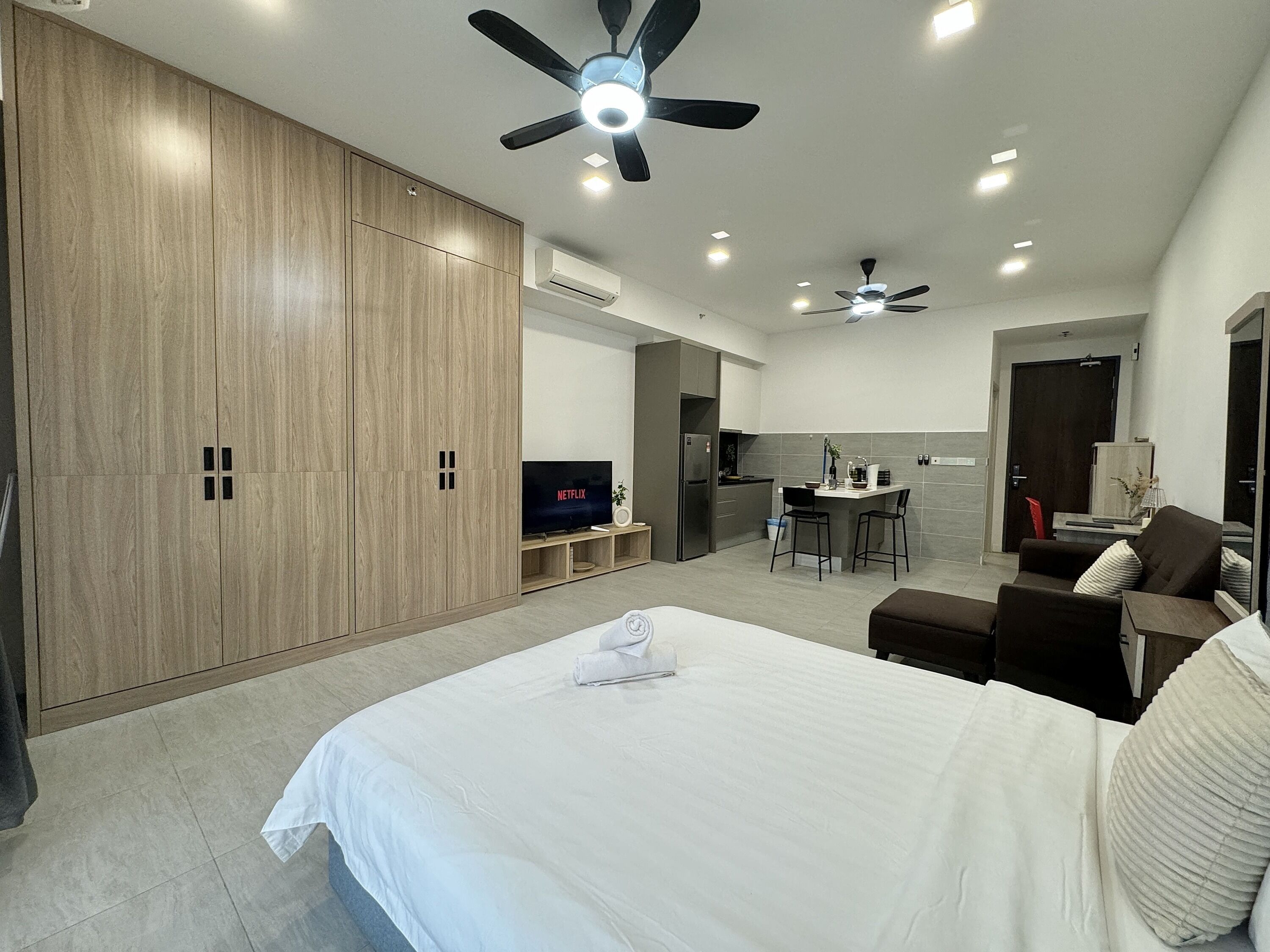 A modern hotel room with a large bed, a TV, a kitchenette, and a ceiling fan.