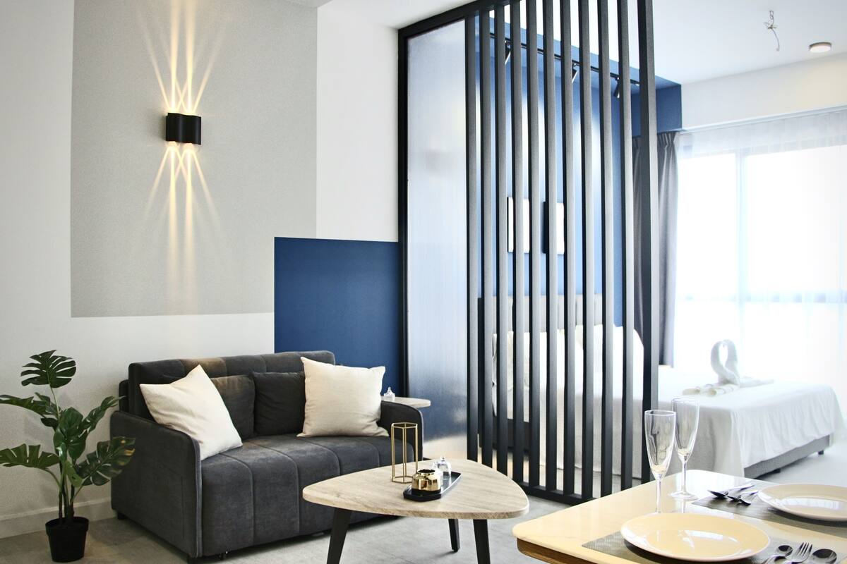 Moderni hotellihuone, jossa on harmaa sohva, pyöreä sohvapöytä tarjottimella, kahdelle katettu ruokapöytä ja valkoisella päiväpeitteellä varustettu sänky.