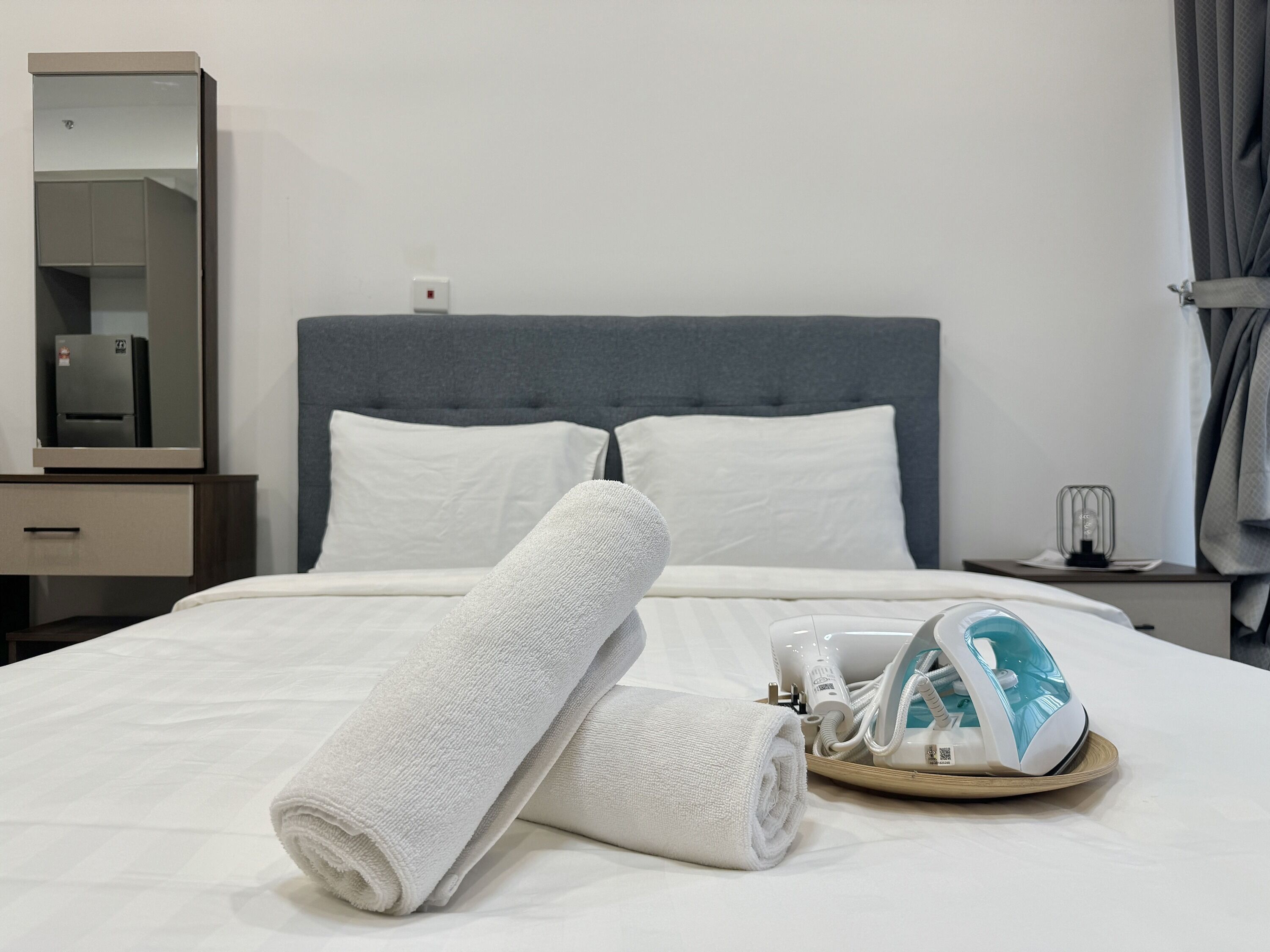 Un lit bien fait, avec du linge de lit blanc, une tête de lit grise et une table de chevet sur laquelle se trouvent une lampe et un plateau avec un sèche-cheveux.