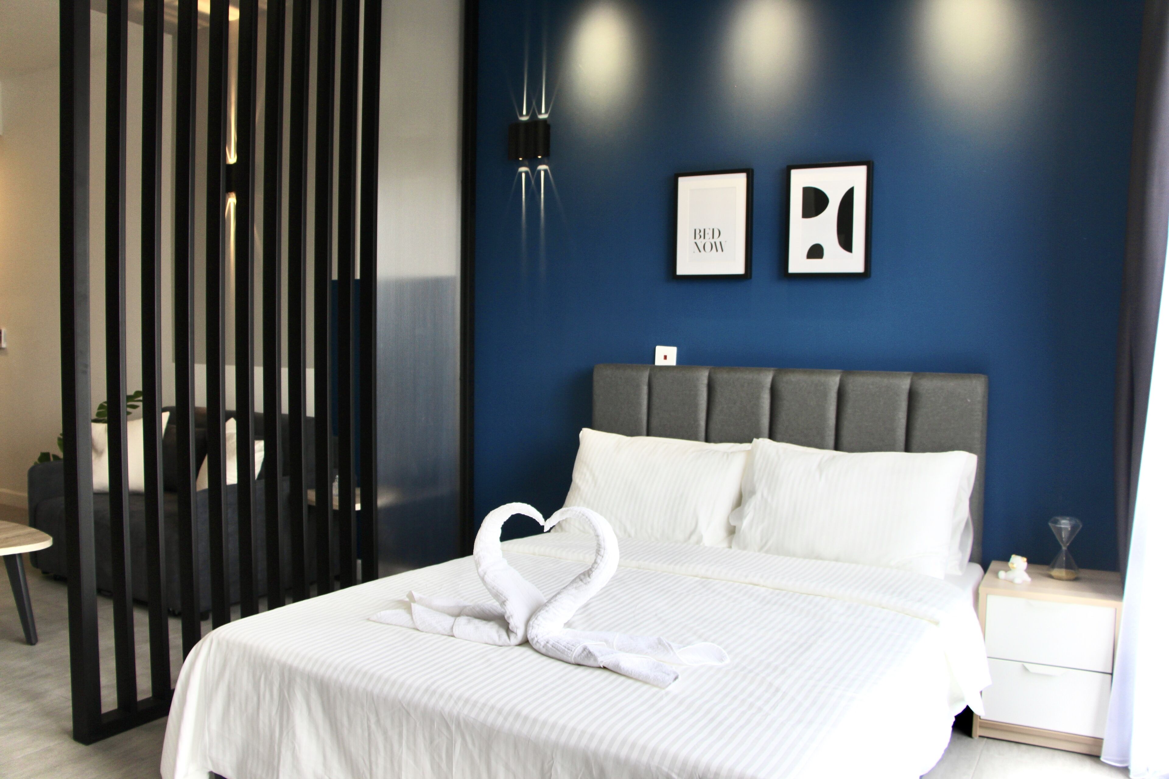 A neatly made bed with a heart-shaped towel arrangement, a grey headboard, and two framed pictures on the wall.