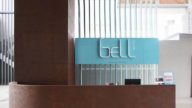 A reception desk with a sign reading "GELL SUITES" and "Sunsurya City" in a modern lobby with a curved brown reception area and vertical light-colored panels.