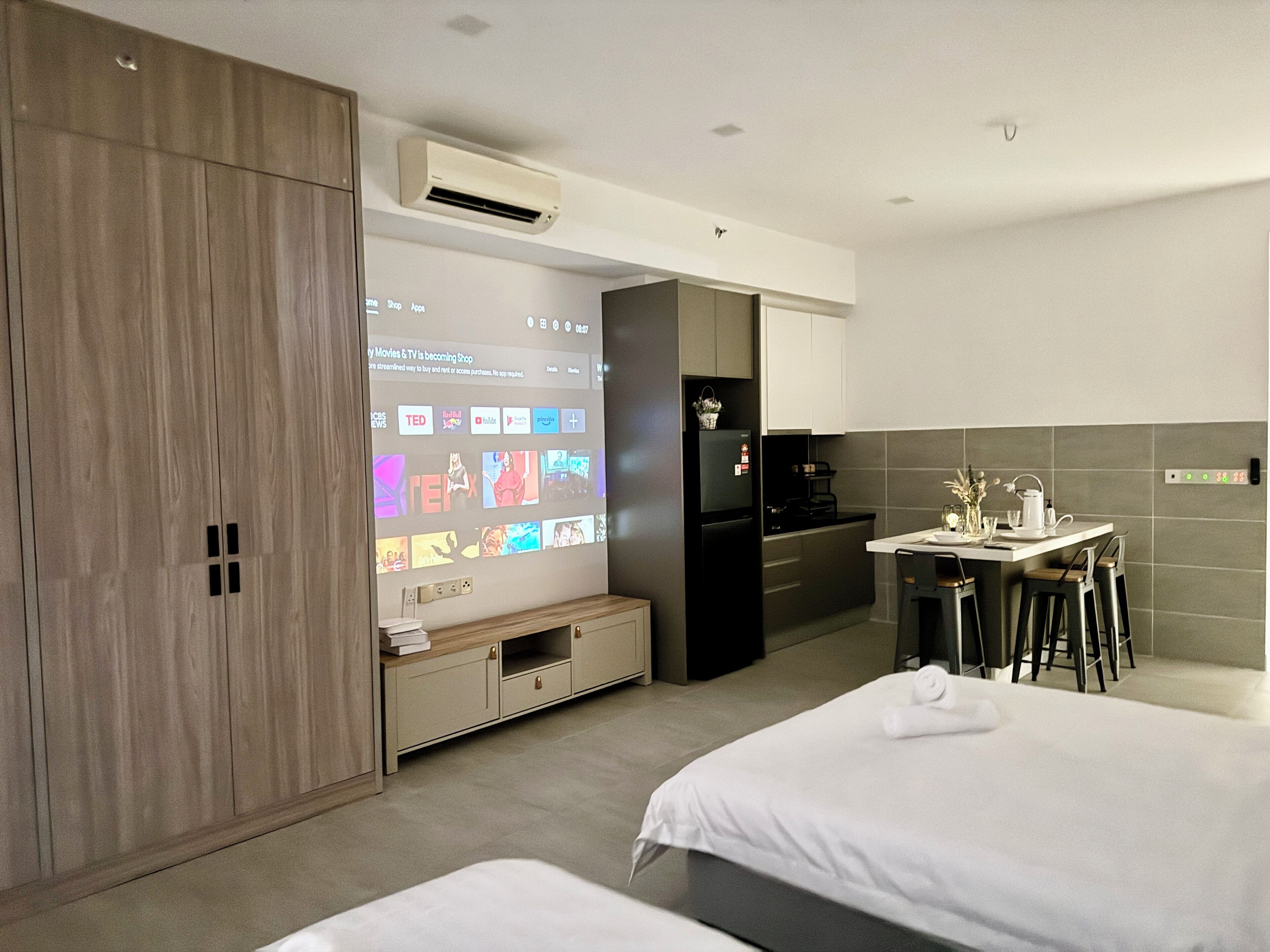 Habitación de hotel moderna con cama, escritorio con silla, kitchenette y televisor mostrando una película y una guía de televisión.