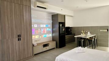 Habitación de hotel moderna con cama, escritorio con silla, kitchenette y televisor mostrando una película y una guía de televisión.