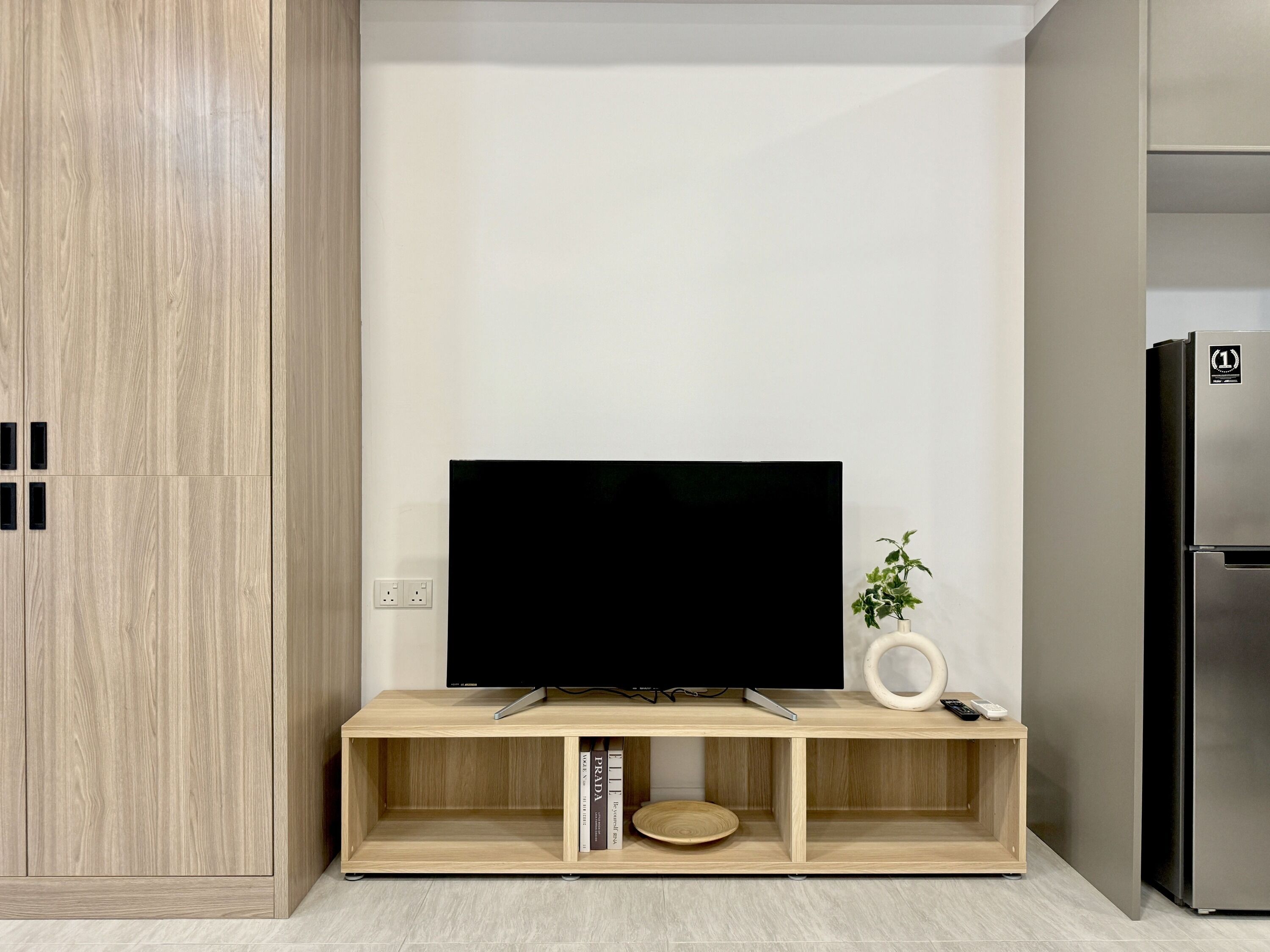 A modern kitchen with a built-in TV on a wooden stand, a refrigerator, and a small plant.