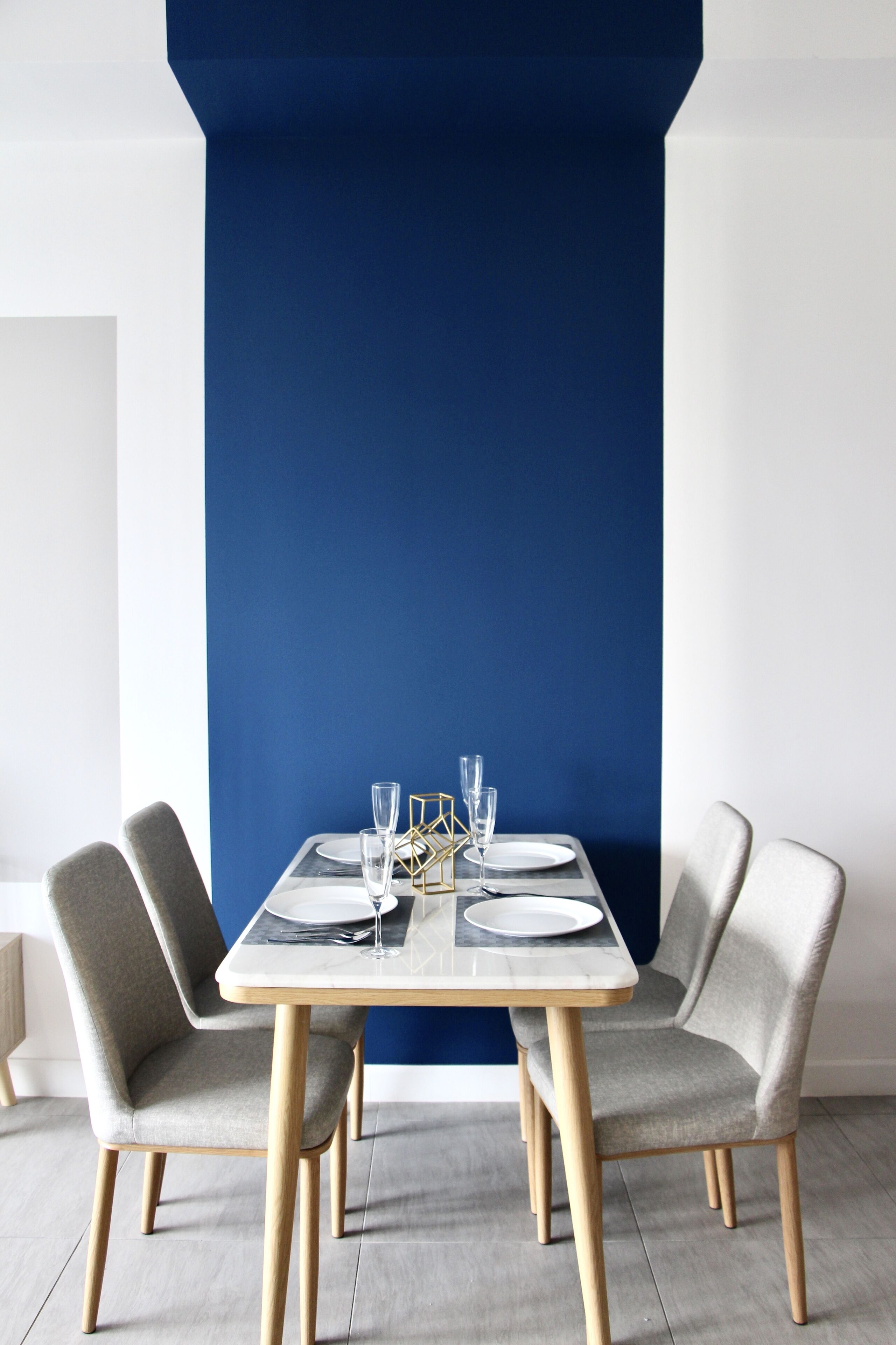 A dining table set with plates, glasses, and cutlery, flanked by two upholstered chairs, against a blue accent wall.