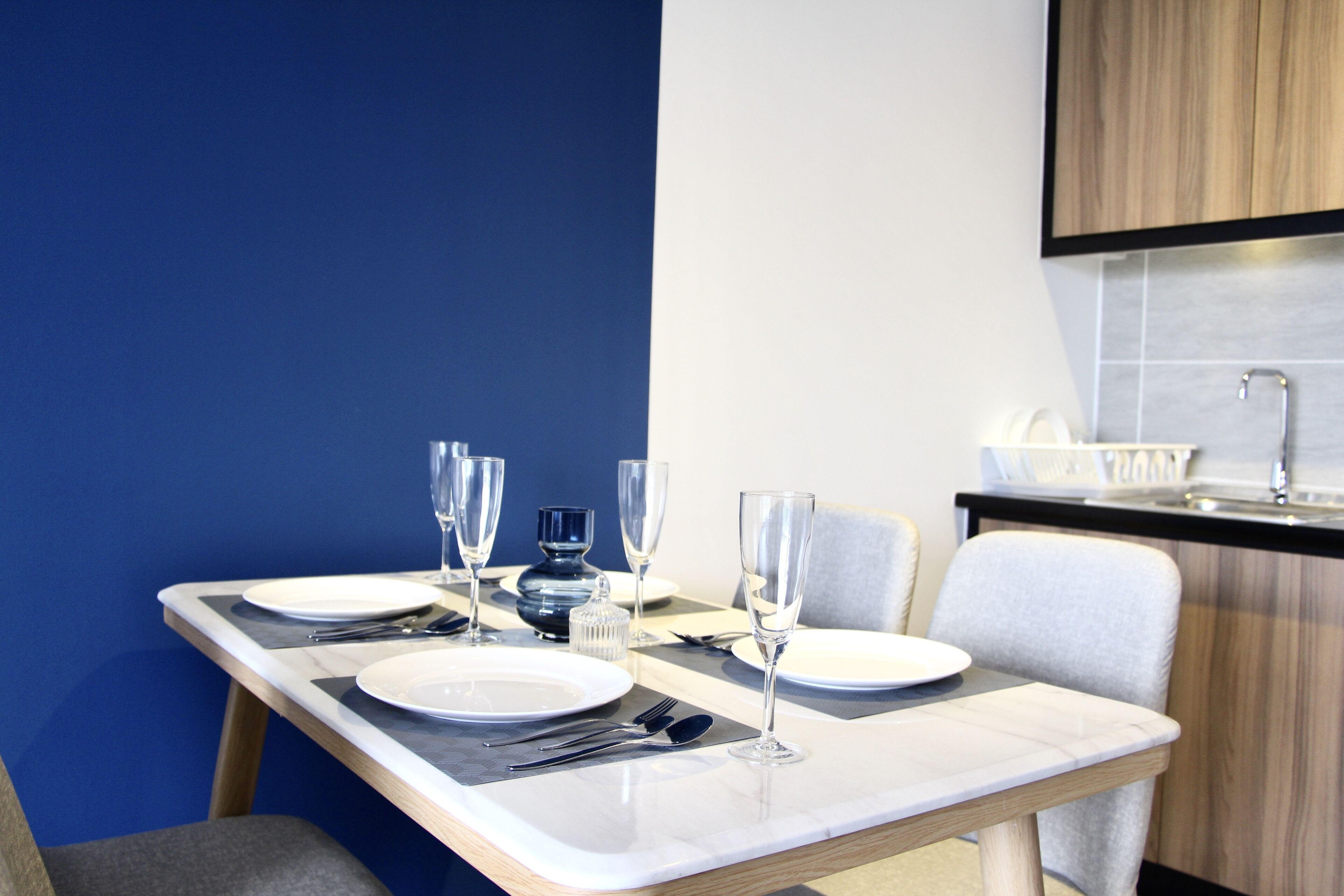 Una mesa de comedor dispuesta con platos, cubiertos y vasos, con una cocina moderna al fondo.