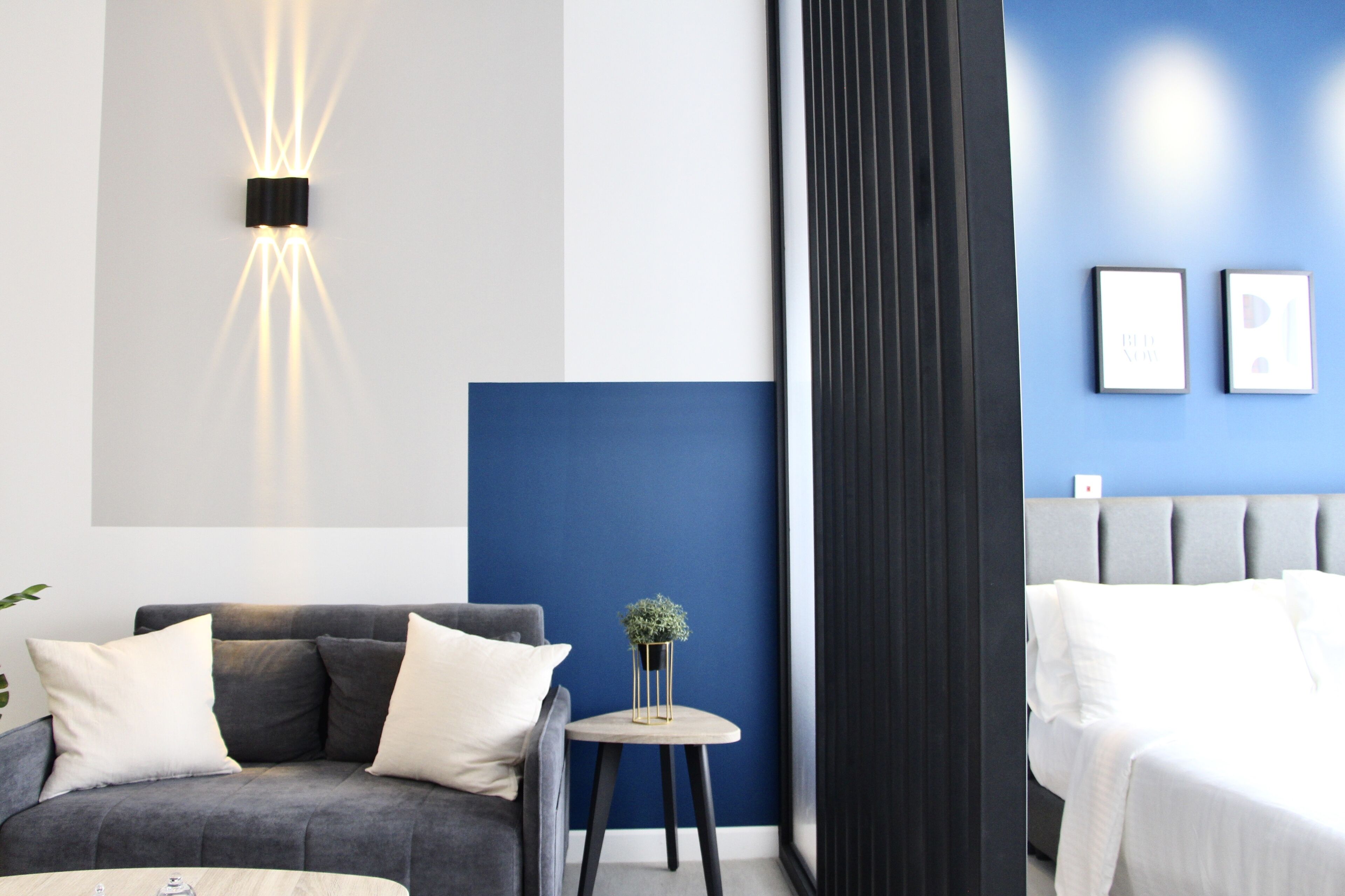 A modern hotel room with a grey sofa, a round side table with a plant, a bed with white linens, and a blue accent wall.