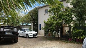 Una entrada residencial con un coche blanco estacionado a la izquierda y un SUV negro a la derecha. Hay plantas en macetas y un acceso cerrado con portón.