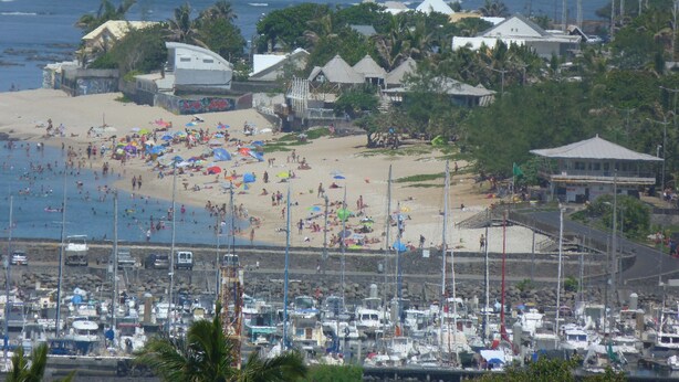 Una playa con muchos sombrillas, un puerto deportivo con barcos y edificios con techos de paja.