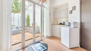 A compact kitchen with a white refrigerator, microwave, and cabinets, a small table with a magazine, and a sliding glass door leading to a patio with plants and a staircase. - MaisonMars Studio 3 Blancarde (Marseille)