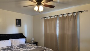 Un dormitorio con cama, ventilador de techo, mesita de noche, lámpara y ventana con cortinas.