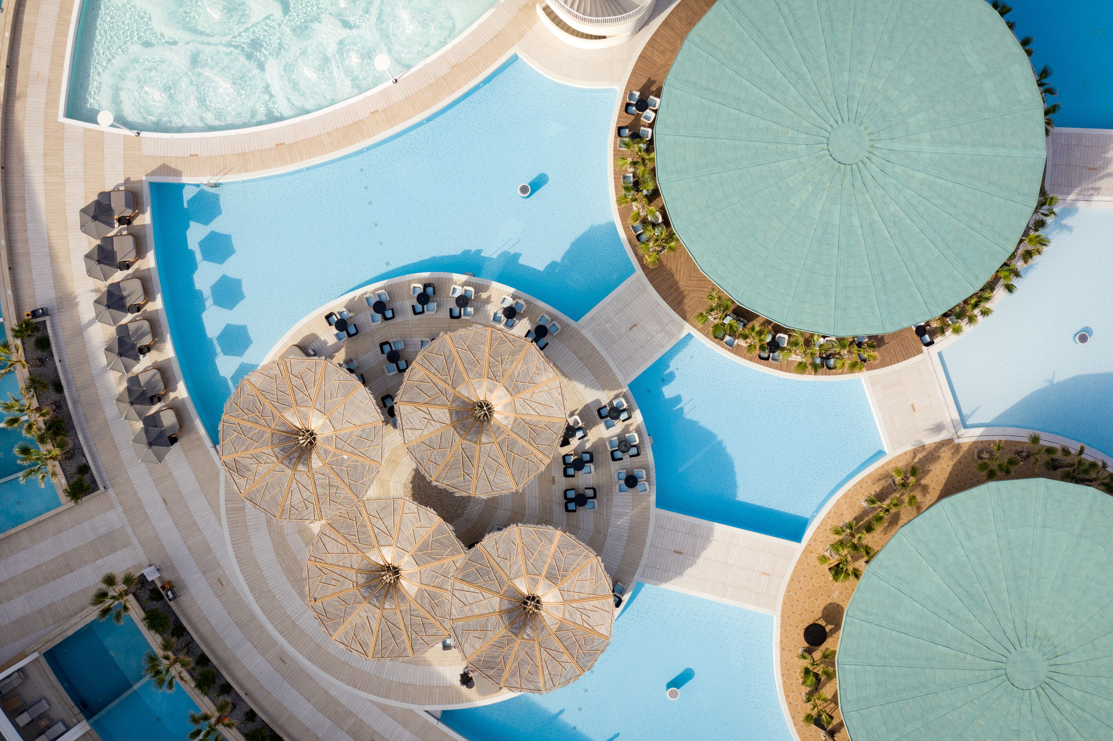 An aerial view of a resort with multiple swimming pools, sun umbrellas, and lounge areas.