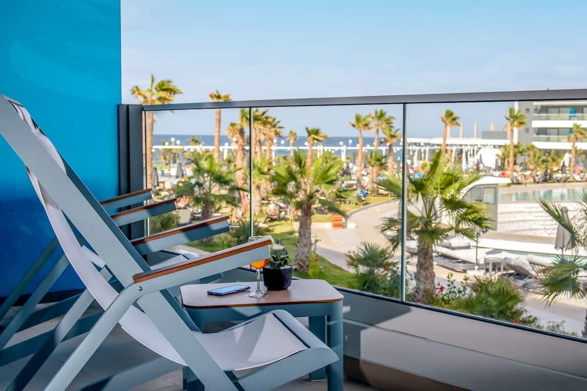 A balcony with two lounge chairs, a small table with a phone and a glass, and a view of a resort area with palm trees and a pool.