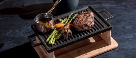 A portable grill with a steak and vegetables on a wooden stand.